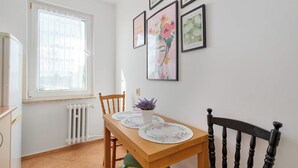 En matplats med ett träbord, stolar, ett fönster med en blommig gardin och konstverk som hänger på väggen.