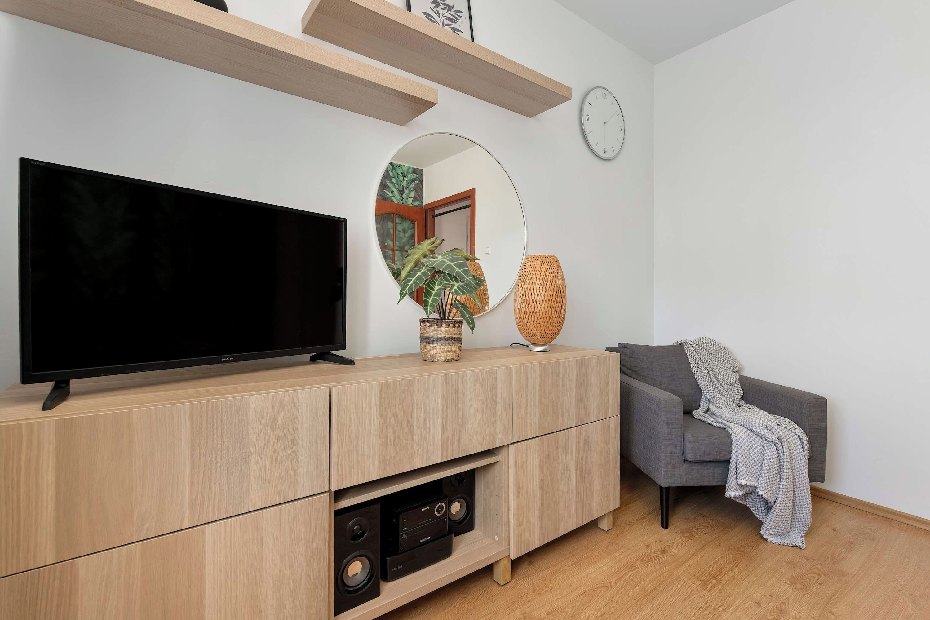 A modern living room with a flat-screen TV on a wooden console, a grey sofa, a woven lamp, and a wall-mounted clock.