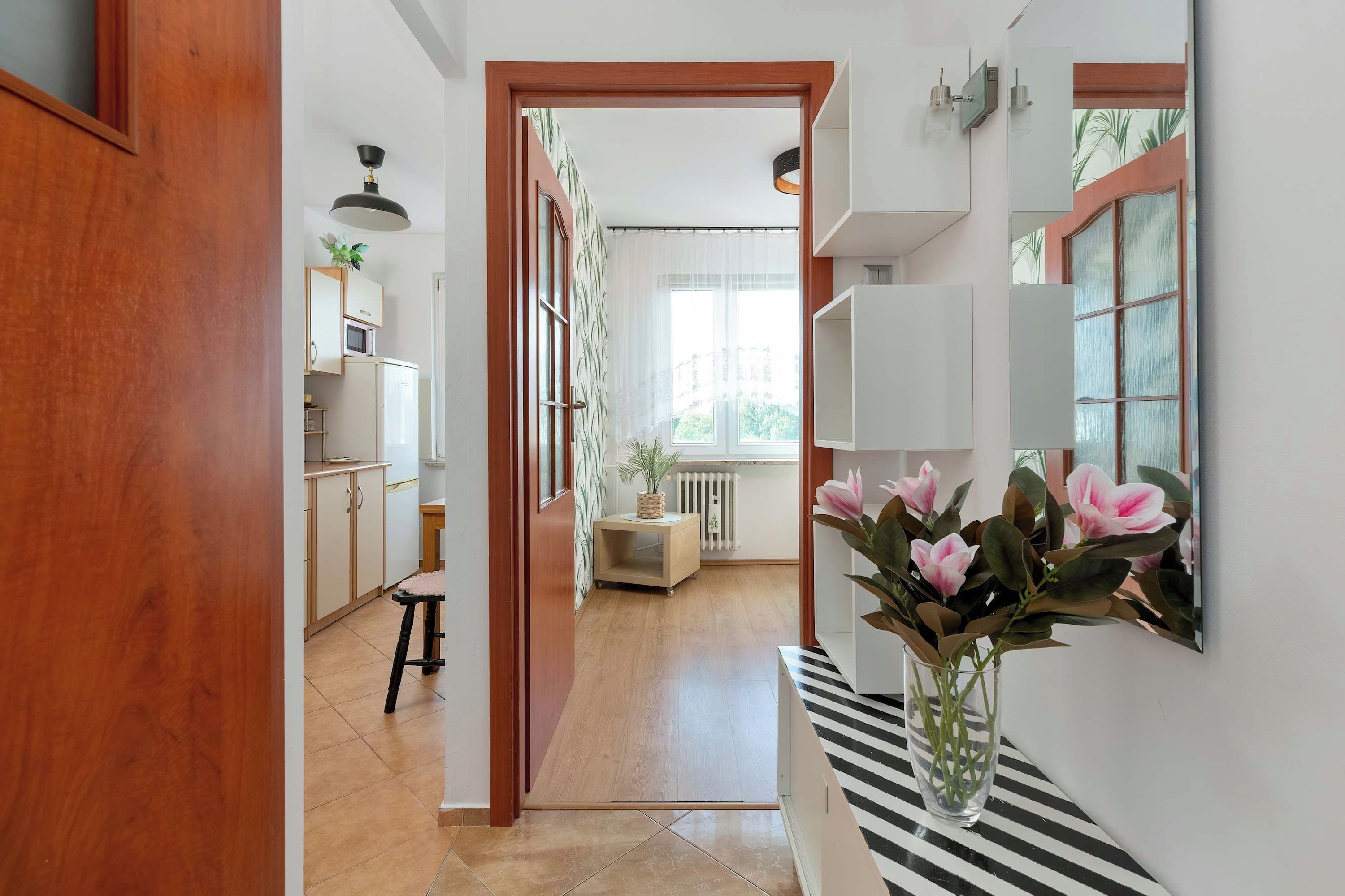 Un pasillo con una puerta de madera, una zona de cocina con armarios y un microondas, un taburete pequeño y una planta en maceta.