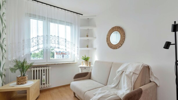 Ett modernt vardagsrum med en soffa, ett litet bord med en växt, ett element och ett fönster med tunna, genomskinliga gardiner.