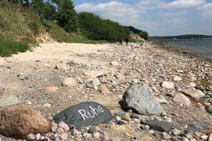 Ein Kiesstrand mit einem Felsen, auf dem 'Ruhe' steht, und einer entfernten Küstenlinie.