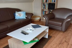 A living room with a brown leather sofa, a white coffee table with remote controls, a green book, and a bookshelf in the background. - Holiday apartment with 3 bedrooms (Dagebüll-Hafen)