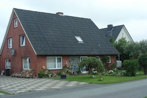 A two-story house with a sloped roof, a visible satellite dish, and a front yard with a checkered pavement and potted plants. - Holiday apartment with 3 bedrooms (Dagebüll-Hafen)