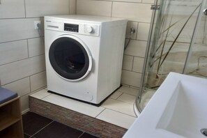 Eine kompakte, weiße Waschmaschine in einem kleinen Waschraum mit einer Glasdusche.