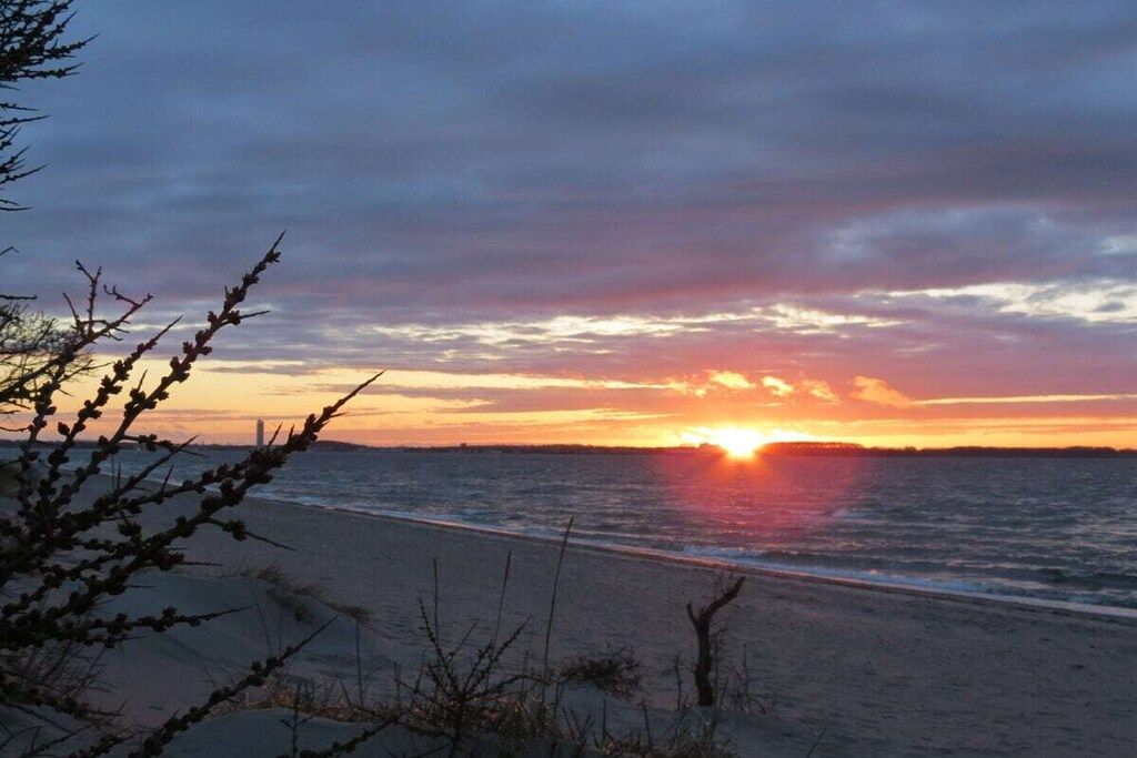 A beach with the sun setting over the horizon, clear skies, and calm sea.