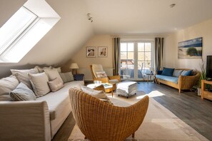 Ein geräumiges Wohnzimmer mit hoher Decke, einer Sofagarnitur, einem Korbsessel und Blick durch ein großes Fenster.