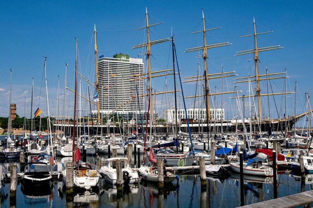 Un port de plaisance où sont amarrés de nombreux voiliers, un grand bâtiment en arrière-plan et un ciel dégagé.