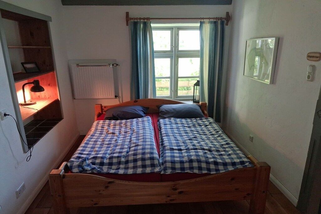 Uma cama de solteiro com estrutura de madeira, mesa de cabeceira com abajur, janela com cortinas e radiador.