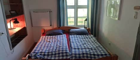 A single bed with a wooden frame, a bedside table with a lamp, a window with curtains, and a radiator.