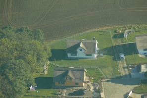 Uma área rural com duas casas, uma com telhado de palha e outra com telhado em duas águas, cercada por campos e uma pequena área isolada por cerca.