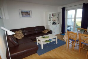 A living room with a brown leather sofa, a white cabinet, a dining table with chairs, and a large window with curtains. - Holiday apartment with 2 bedrooms (Schönberger Strand)