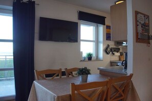 A dining area with a table and chairs, a television mounted on the wall, a window with a view, and a kitchen area visible through an open door. - Holiday apartment with 2 bedrooms (Schönberger Strand)