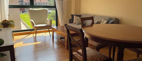 A room with a wooden dining table, chairs, a sofa, and a window with a view of houses and greenery.