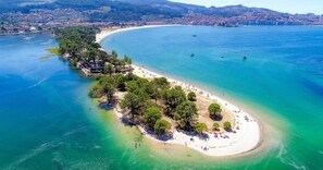 An aerial view of a coastal area with a beach, clear waters, and a small island with trees.