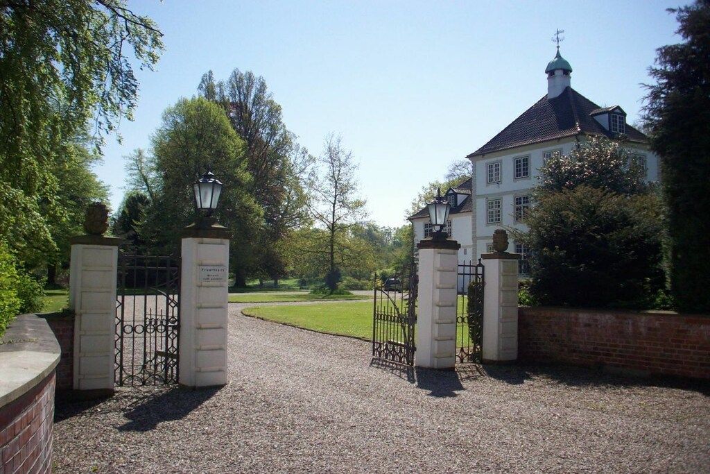 Uma entrada com portão para um edifício de estilo arquitetônico tradicional, cercada por um caminho de cascalho e vegetação.