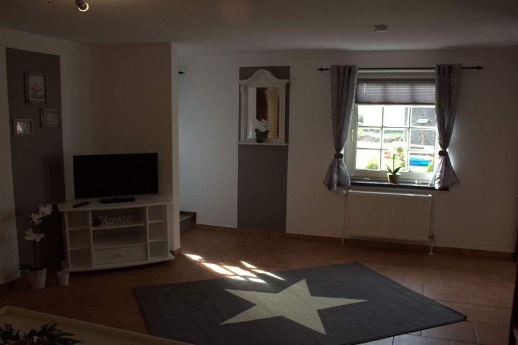 Ein Wohnzimmer mit einem Fernseher auf einem weißen Schrank, einem Teppich mit Sternenmuster, einem Fenster mit Vorhängen und einer Heizung.
