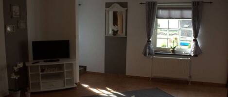 Ein Wohnzimmer mit einem Fernseher auf einem weißen Schrank, einem Teppich mit Sternenmuster, einem Fenster mit Vorhängen und einer Heizung.