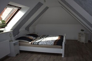 Een slaapkamer met een schuine kap, een bed met beddengoed met een patroon, een nachtkastje met een lamp en een kleine plant.