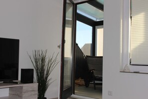 A room with a television on a wooden stand, a potted plant, and a sliding glass door leading to an outdoor area with a chair and a table. - Swallow's nest upper floor (Maasholm)