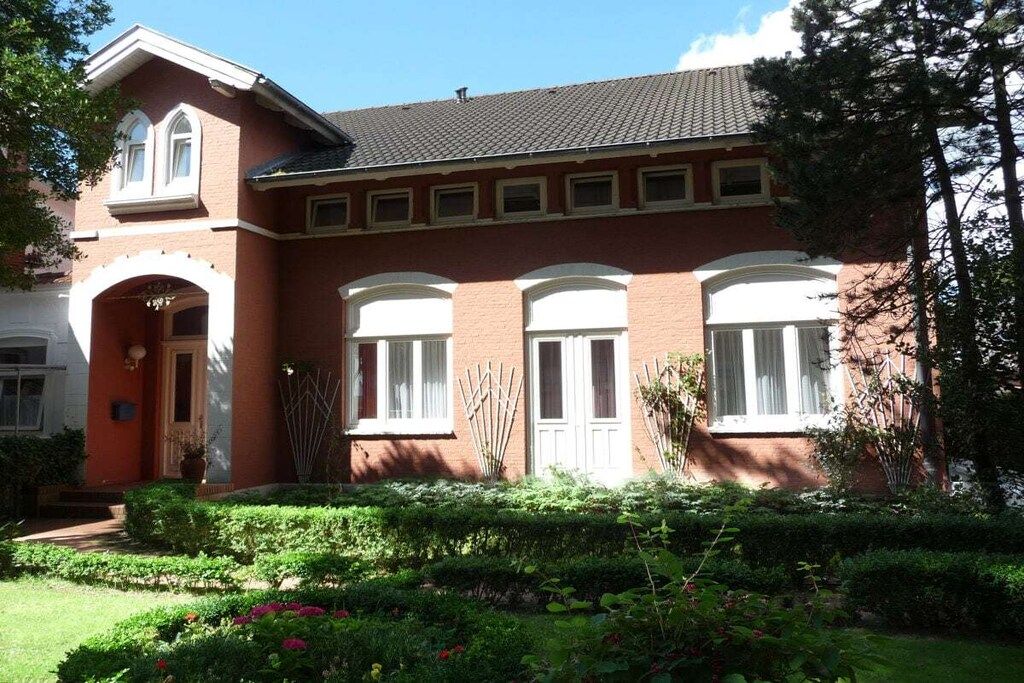Une maison à deux étages, construite en briques, avec une façade symétrique, une entrée centrale dotée d’une porte et de fenêtres, et un jardin bien entretenu où poussent diverses plantes et fleurs.