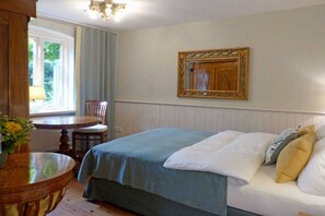 Ein Zimmer mit einem Bett, einem runden Holztisch mit Stuhl, einem Fenster mit Vorhängen und einem gerahmten Spiegel an der Wand.