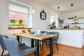A modern kitchen with a wooden table, grey chairs, a clock, and a window with a view of a brick building. - Meerhuus Carolinensiel House 3 (Carolinensiel)