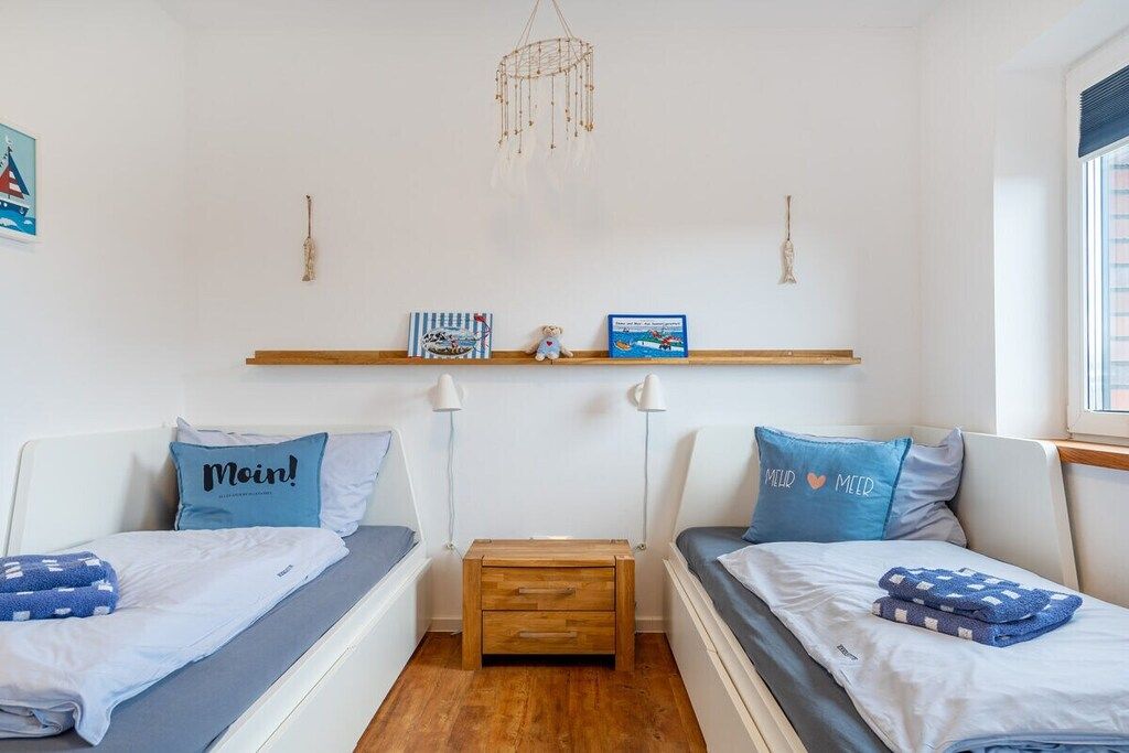 A room with two beds, a wooden nightstand, a wall-mounted shelf with books and a small figurine, and a hanging dreamcatcher.