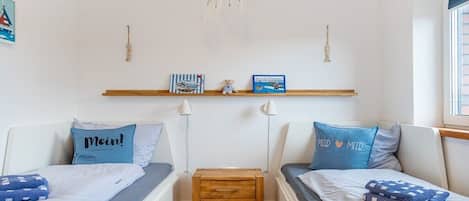 Una habitación con dos camas, una mesita de noche de madera, un estante en la pared con libros y una pequeña figurita, y un atrapasueños colgando.