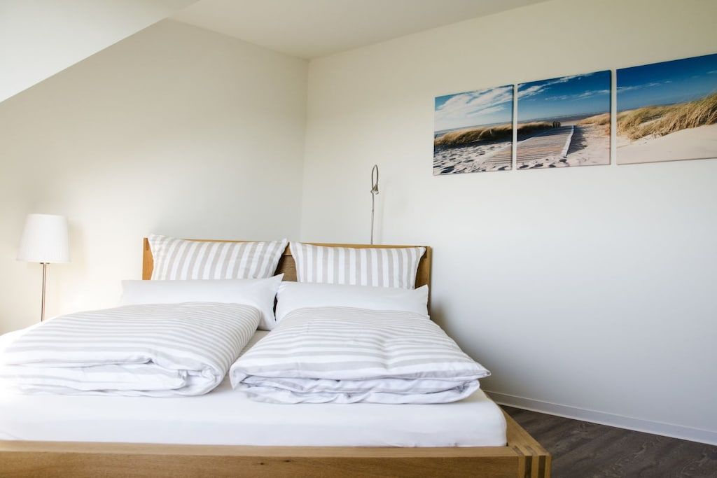 Un lit bien fait, avec une literie blanche et des oreillers à rayures, une lampe de chevet et un mur orné de trois tableaux représentant une scène de plage.
