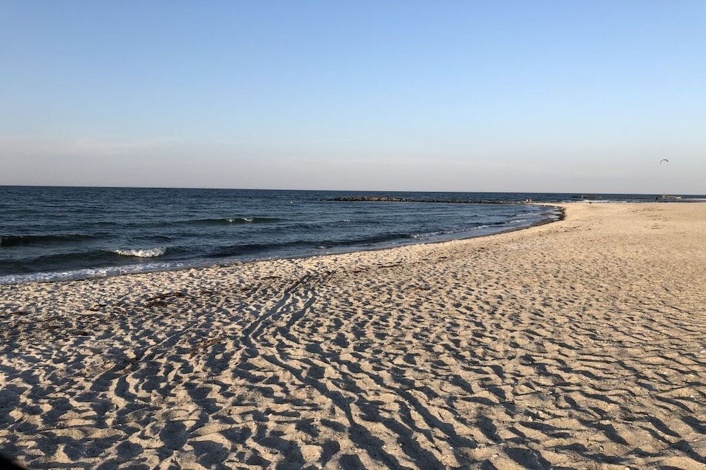 En sandstrand med milda vågor, en klar himmel och en brygga som ligger en bit bort.