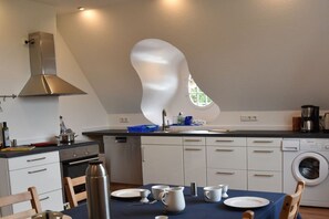 A modern kitchen with a unique ceiling design, a dining table set for four, and a built-in washing machine. - Holiday apartment at the Baltic Sea resort (Flensburg)