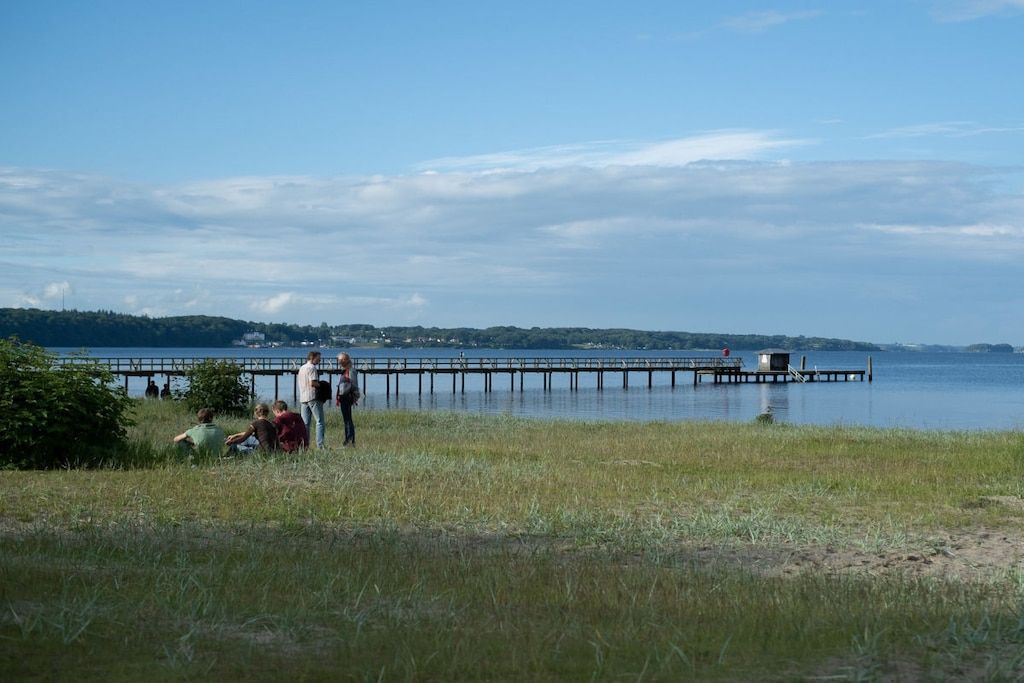 A lakeside view with a dock extending into the water, surrounded by grassy areas and distant hills.