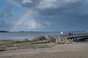 Una spiaggia con un molo di legno che si protende su acque calme, un arcobaleno nel cielo e alcune persone sul molo.