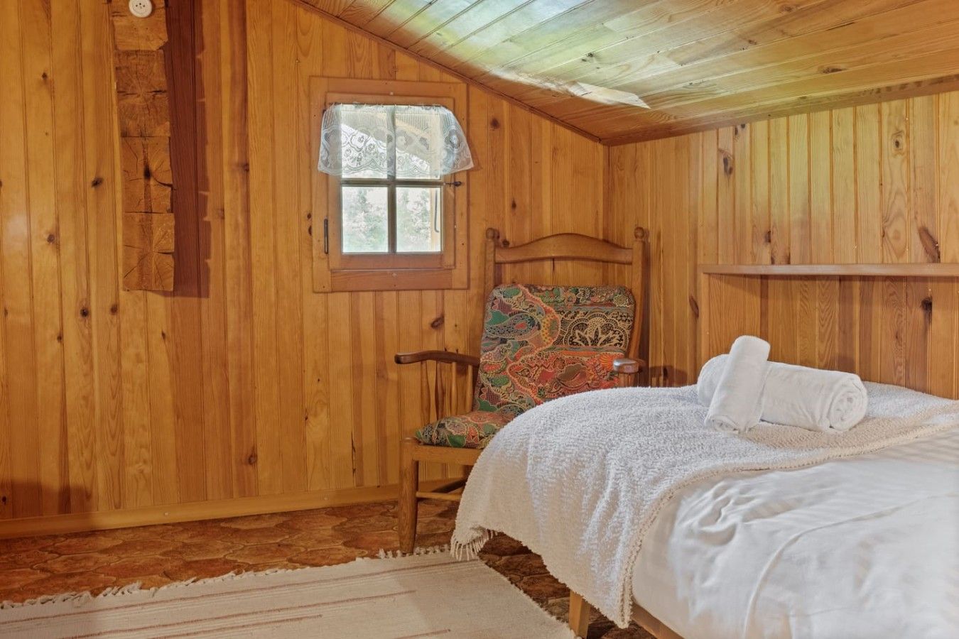 Quarto de madeira com cama, cadeira, janela e uma pequena estante.