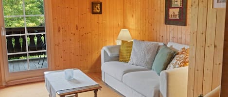 Una habitación acogedora con un revestimiento de madera, un sofá blanco con cojines, una mesita y una ventana con vistas a zonas verdes.