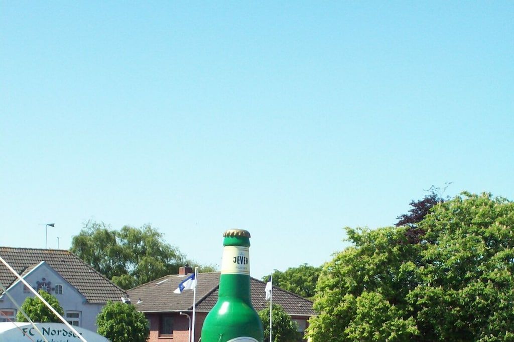 A large green bottle with a label, a house with a blue roof, and a tree in the background.