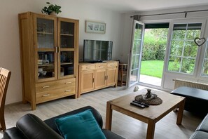 A living room with a wooden cabinet, a television, a coffee table with remote controls, and a view of a garden through a glass door. - Holiday home with 3 bedrooms (Wangerland \/ Hooksiel)