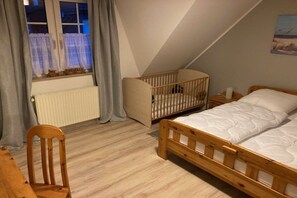 A bedroom with a wooden bed, a crib, a chair, and a window with curtains. - Holiday home with 3 bedrooms (Wangerland)