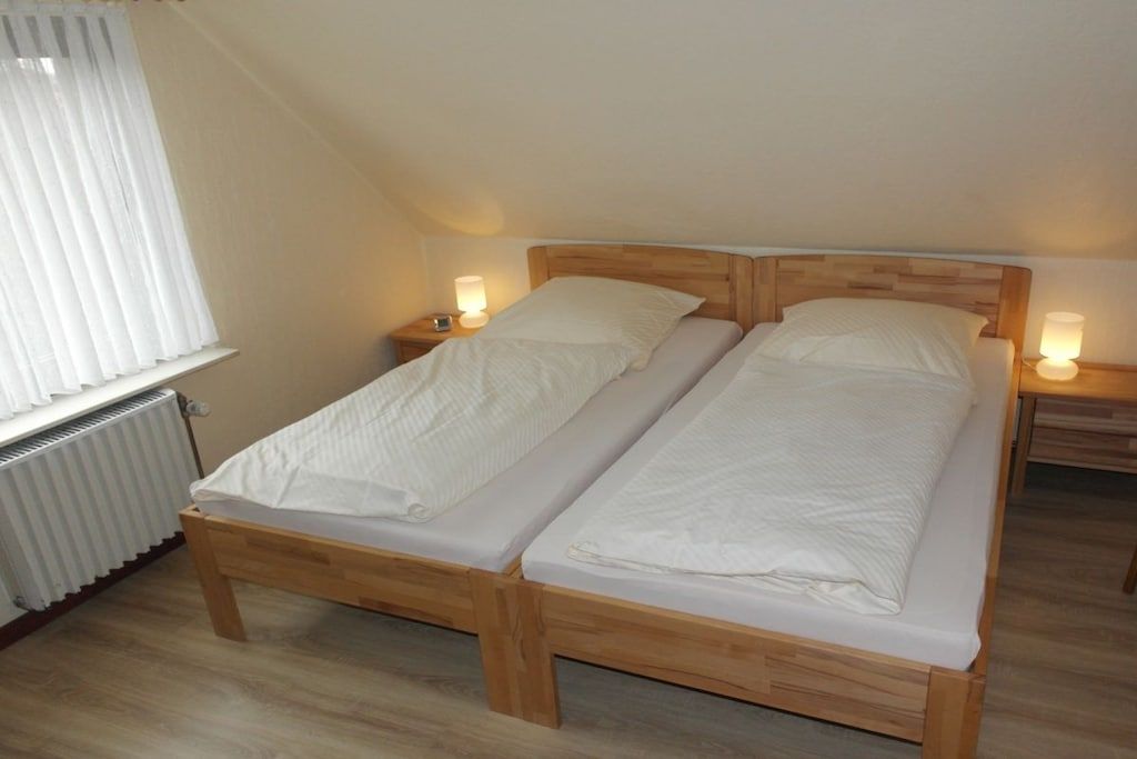 A twin bed with wooden headboard and footboard, two bedside tables with lamps, a radiator, and a window with white curtains.