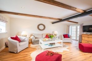 A spacious living room with a white sofa, a red ottoman, a coffee table, and a television. - White Cottage Estate Priesholz (Rabenholz)