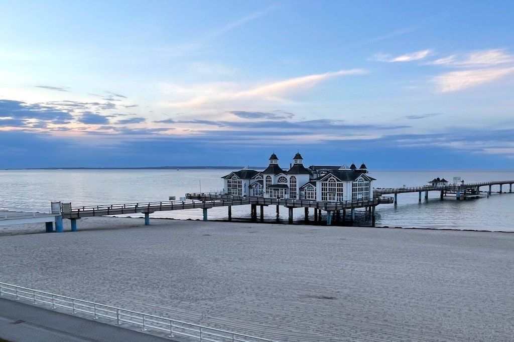 Un edificio costero con un muelle que se adentra en el agua, una playa al frente y un cielo despejado.