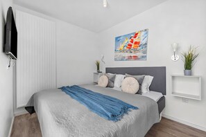 A modern bedroom with a bed, a painting, a plant, and a TV mounted on the wall. - Kielwasser – Prime Location, Stunning Sea Views, Steam Shower & Private Parking (Wendtorf)