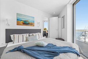 A modern bedroom with a large bed, a painting of a beach scene, and a view of a marina through the window. - Kielwasser – Prime Location, Stunning Sea Views, Steam Shower & Private Parking (Wendtorf)