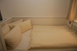 A single bed with a mattress and pillows in a room with white walls and a window. - House Oland Holiday Apartment 5 (Wenningstedt)