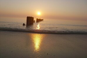 Une scène de plage où le soleil se couche sur l’océan, créant un reflet sur le sable mouillé.