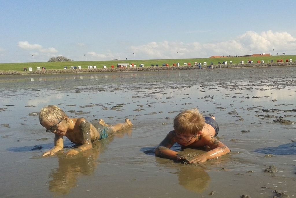 Dos personas en una masa de agua turbia, posiblemente una playa o río, con un área verde y construcciones al fondo.