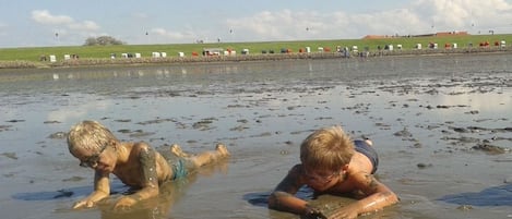 Dos personas en una masa de agua turbia, posiblemente una playa o río, con un área verde y construcciones al fondo.