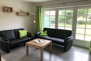 Ein Wohnzimmer mit zwei schwarzen Ledersofas, einem Holztisch und einem großen Fenster mit Blick auf Grünflächen.