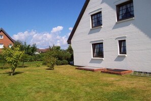 Un bâtiment blanc à deux étages, doté d’un toit plat, d’un petit jardin devant et surplombé d’un ciel bleu clair.