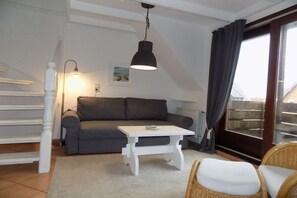 Ein gemütliches Wohnzimmer mit einem grauen Sofa, einem weißen Couchtisch, Korbstühlen, einer weißen Treppe und einem großen Fenster mit Vorhängen.